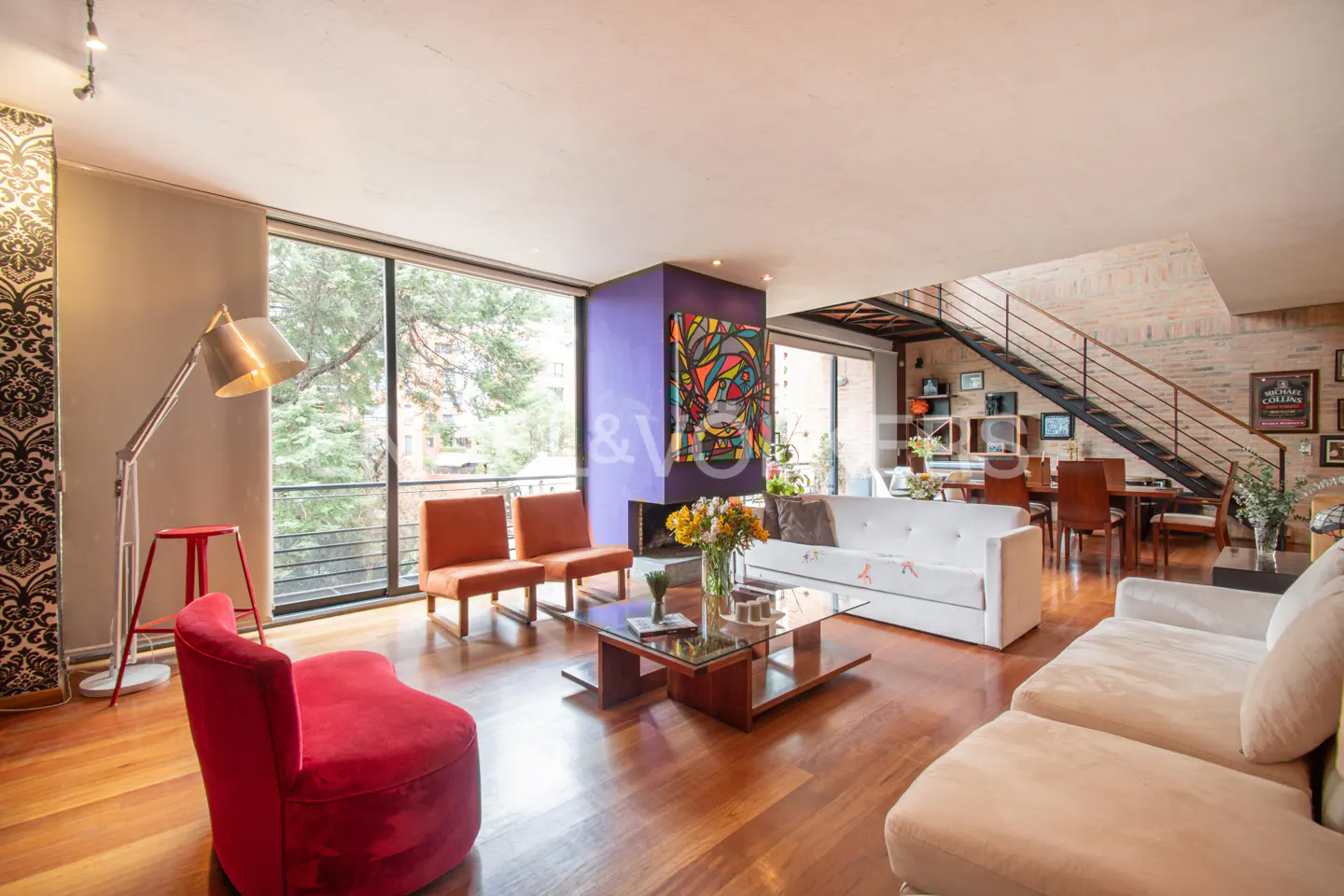 Bright living room with hardwood floors, sofas, chairs, and a fireplace. A staircase leads to the second floor. Large windows offer a view of trees.