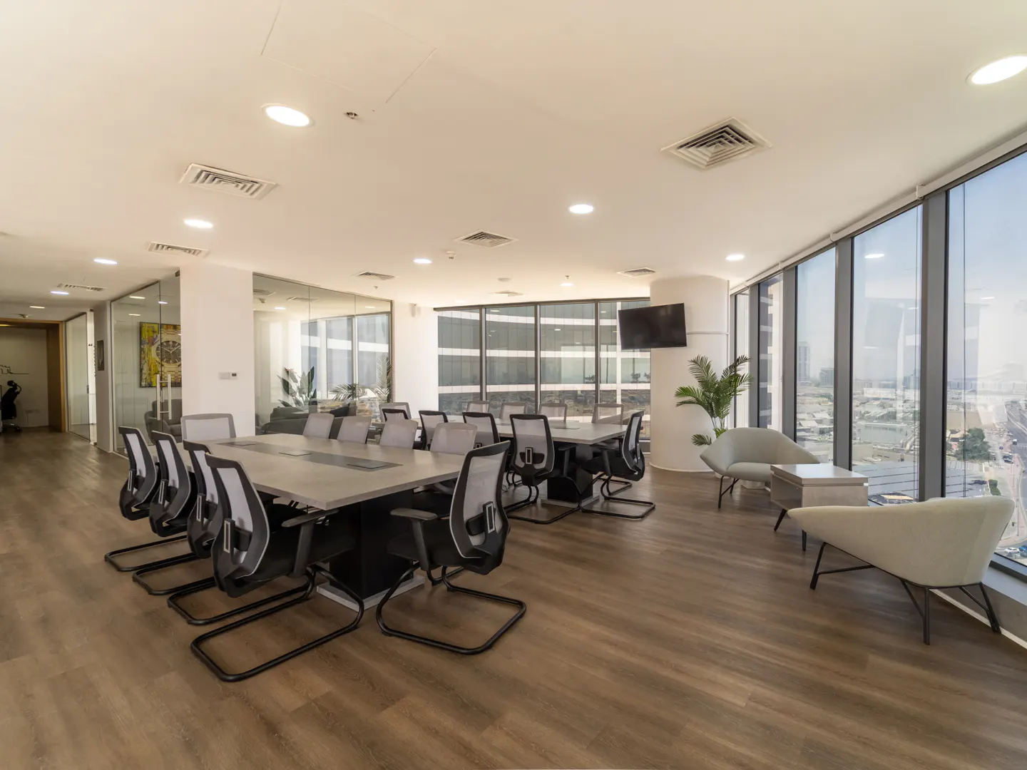 Bright office space with a long conference table, chairs, and lounge area with large windows overlooking a city.