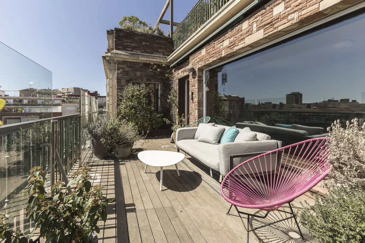 Outdoor patio with wood flooring, gray sofa with blue pillows, pink chair, white table, and potted plants. Brick building and city view in background.