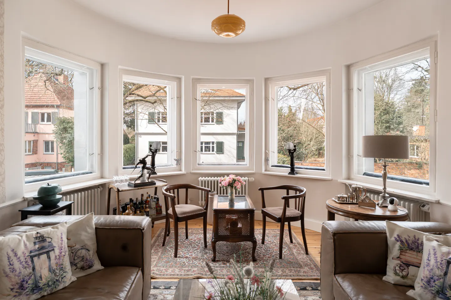 Bright living room with three large windows, brown leather couches, wooden chairs, and a patterned rug. Outside are houses and trees.