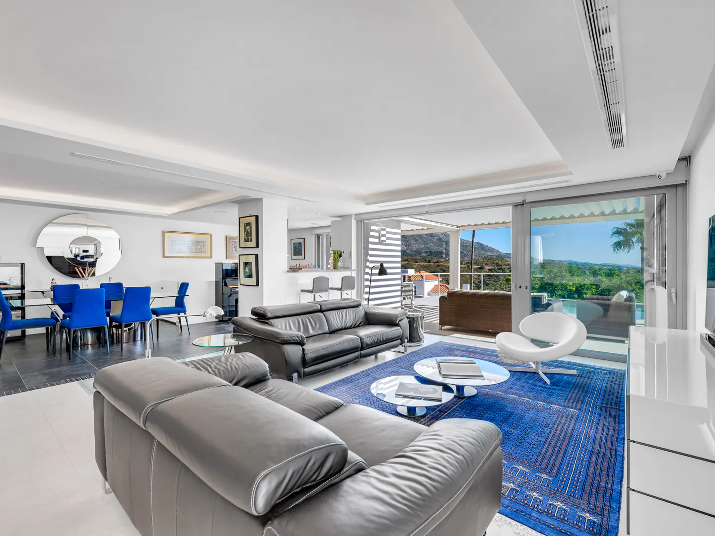 Bright, modern living room with black leather sofas, blue rug, and dining area with blue chairs. Sliding glass doors open to a balcony with mountain views.