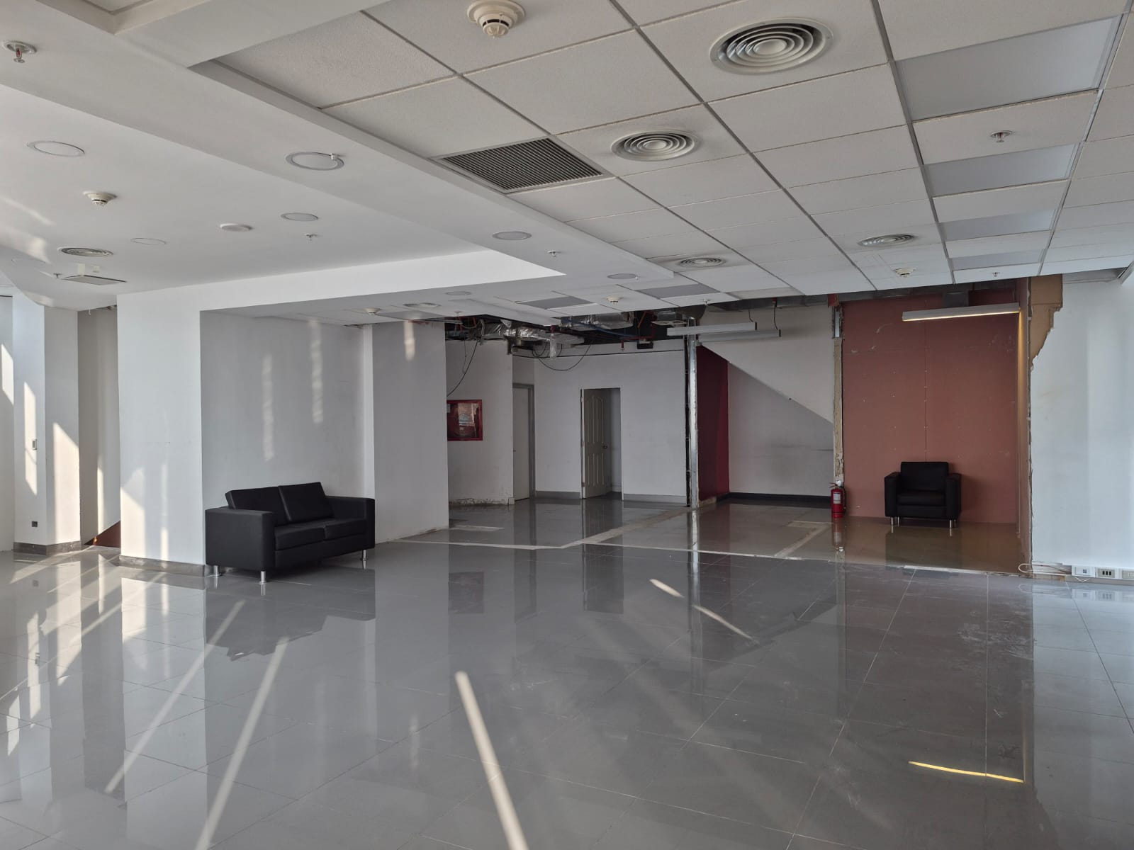 Empty office space with gray tile floor, white walls, and a black sofa. Ceiling has square tiles and vents.