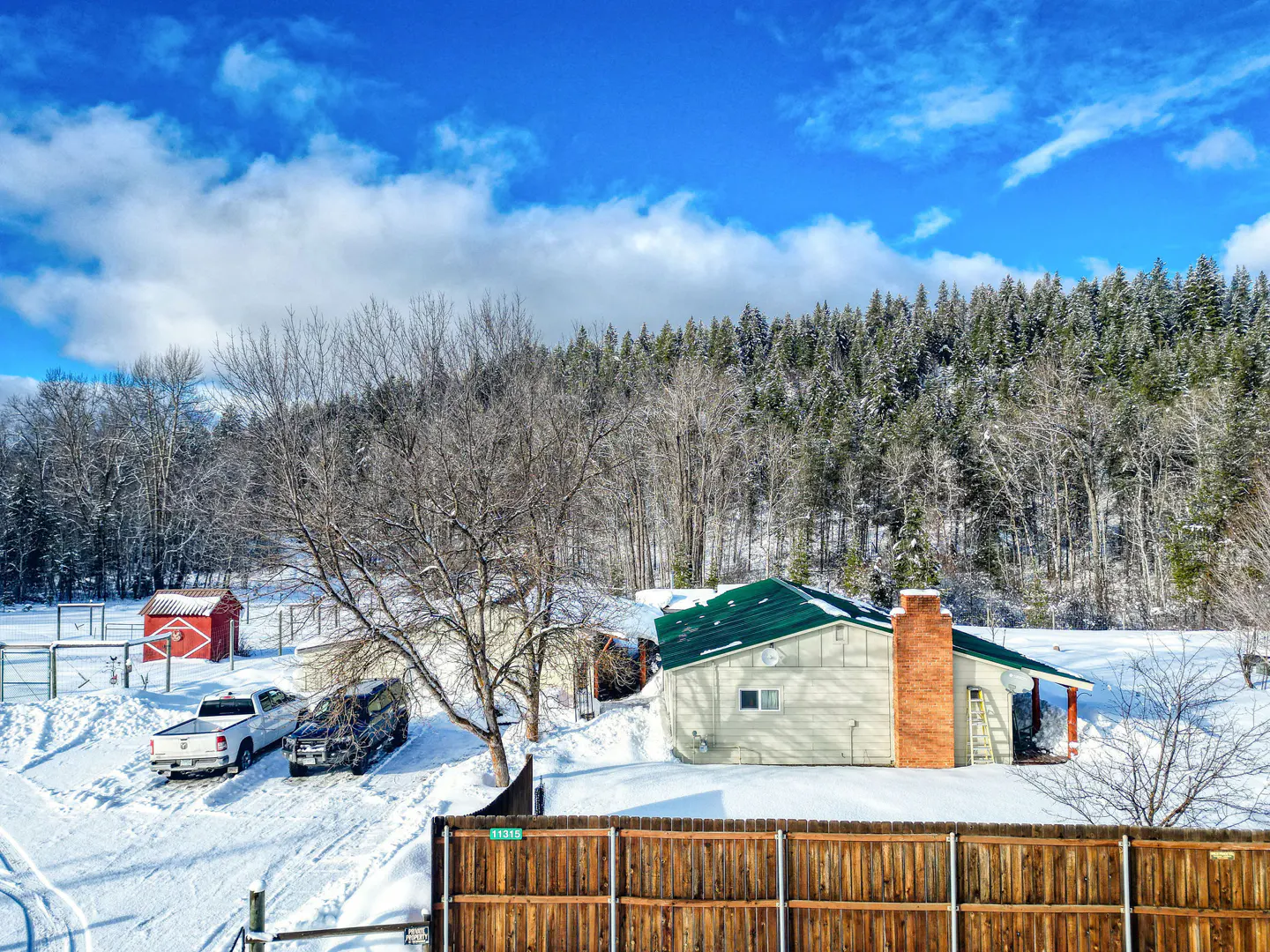 Snowy landscape with a house, red shed, and vehicles. A wooden fence is in the foreground, with a forest backdrop under a blue sky.