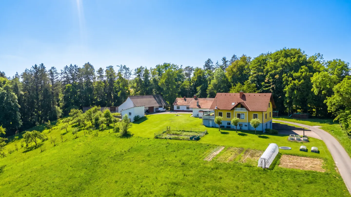 Aerial view of a yellow house with a red roof, surrounded by green grass, trees, and a blue sky. A garden is in the foreground.