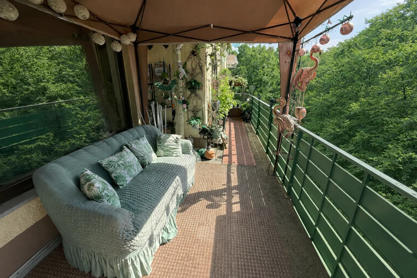 Cozy balcony with a green couch, pillows, plants, and flamingo decor overlooking lush green trees.