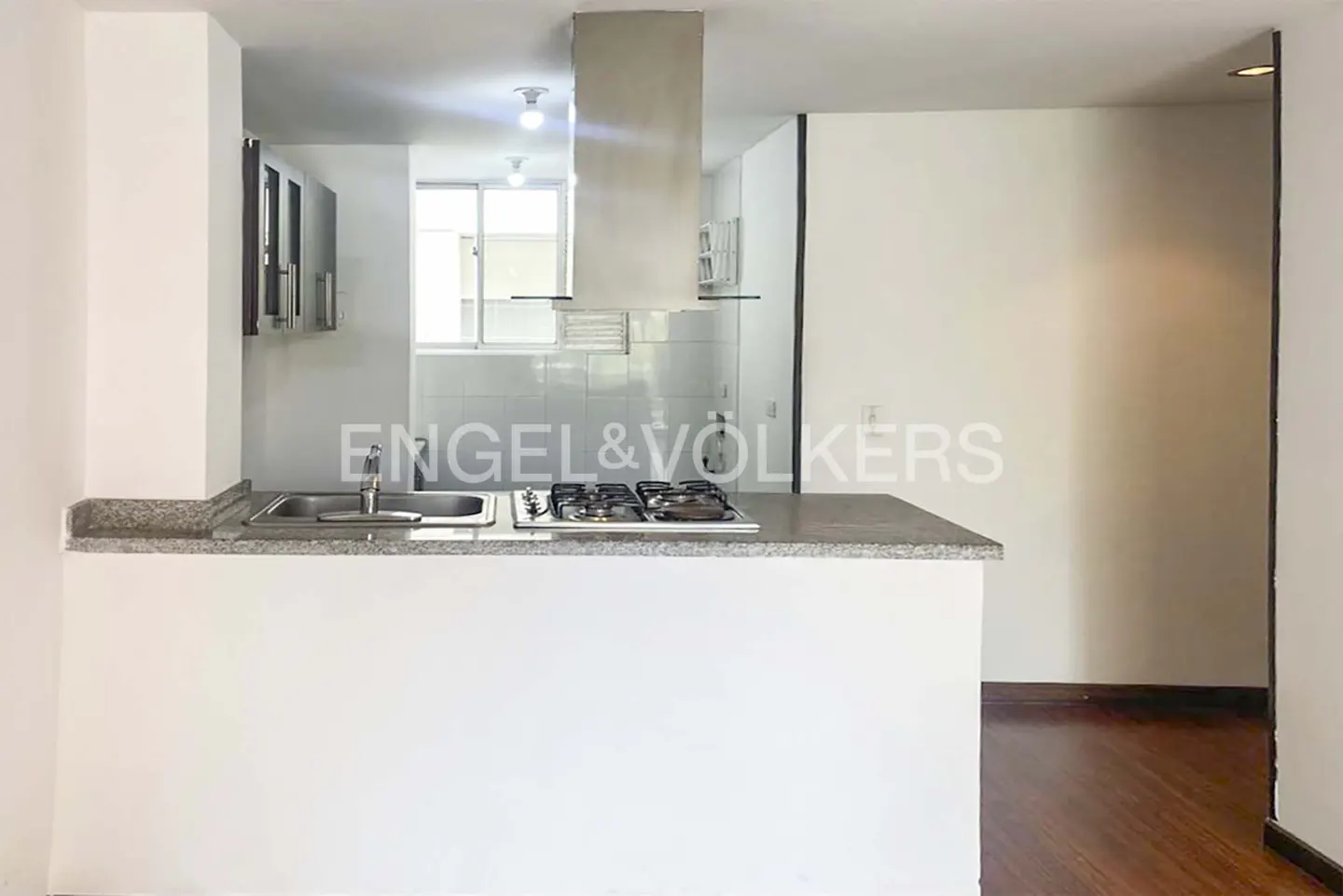 A bright kitchen with a sink, gas stove, and stainless steel range hood. The walls are white, and the floor is wood.