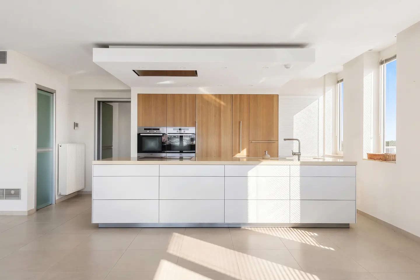 Bright, modern kitchen with white cabinets, wood accents, and stainless steel appliances. Sunlight streams through the windows.