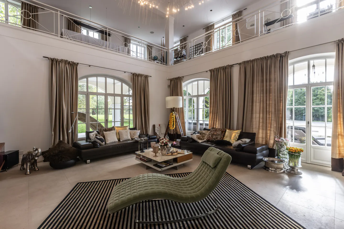 Bright living room with high ceilings, large windows, and a mezzanine. Two black sofas, a green chaise lounge, and a striped rug furnish the space.