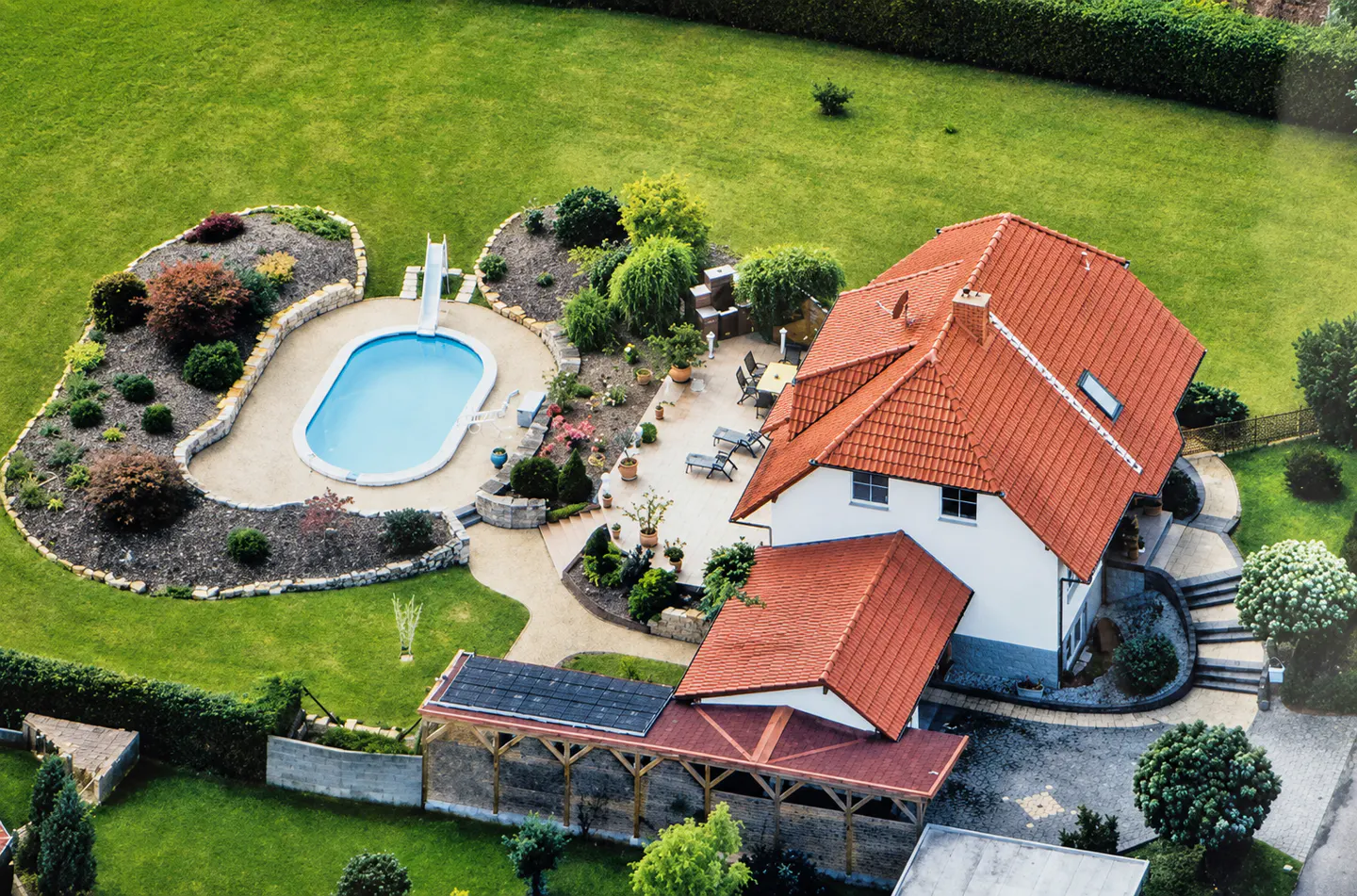Aerial view of a white house with a red tile roof, a swimming pool with a slide, and a large, landscaped green backyard.