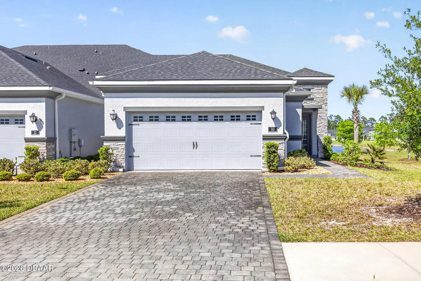 A modern, single-story home with a gray roof, white stucco, stone accents, and a gray brick driveway.