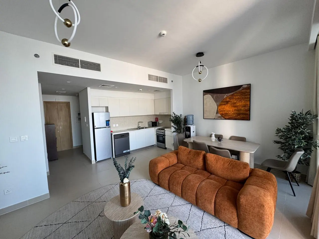 Bright, modern apartment interior with an orange sofa, white kitchen, and dining area. A patterned rug sits under a round coffee table.