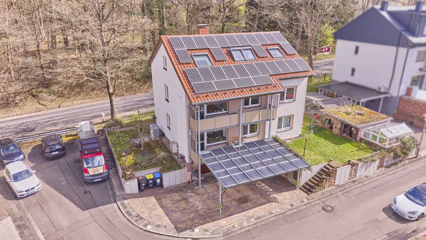 Aerial view of a three-story house with solar panels on the roof and carport. Cars are parked on the street.