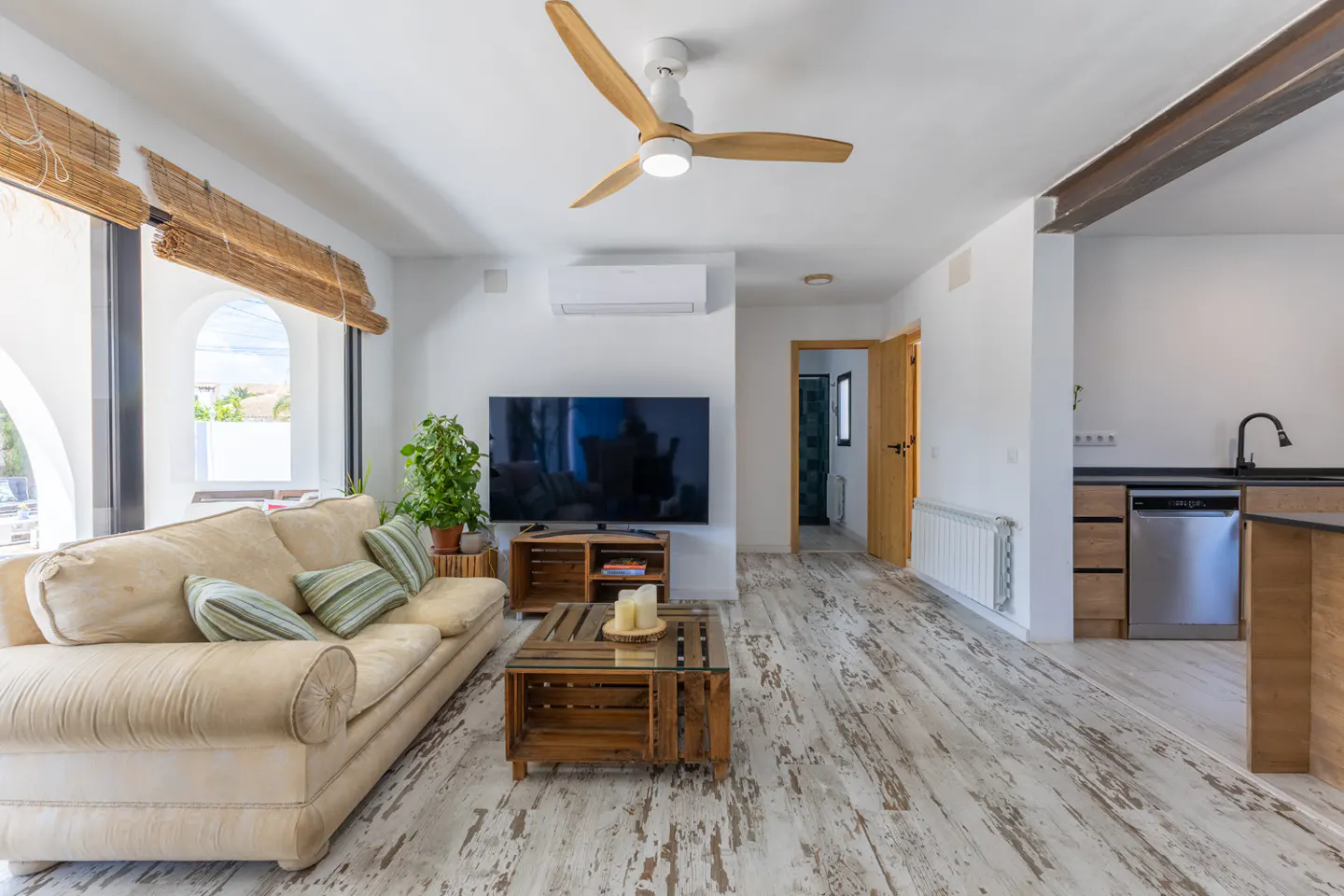 Bright living room with white walls, distressed wood floors, and a beige sofa. A wooden ceiling fan and a TV are also visible.
