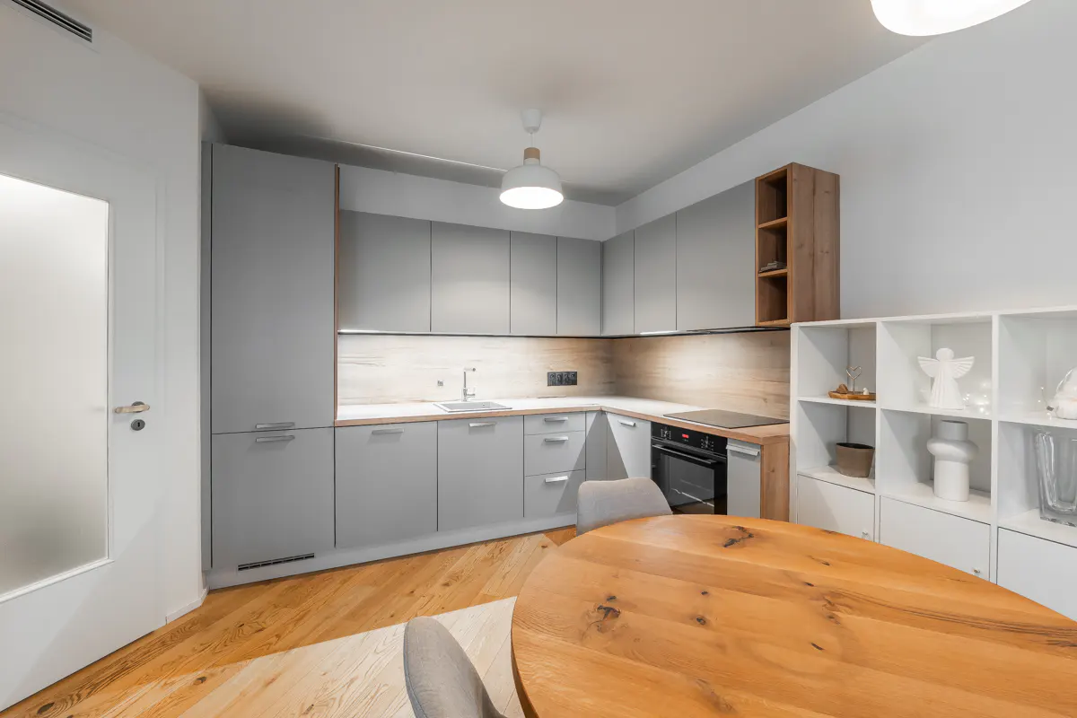 A modern kitchen with gray cabinets, wood countertops, and a round wooden table. White shelves display decorative items.