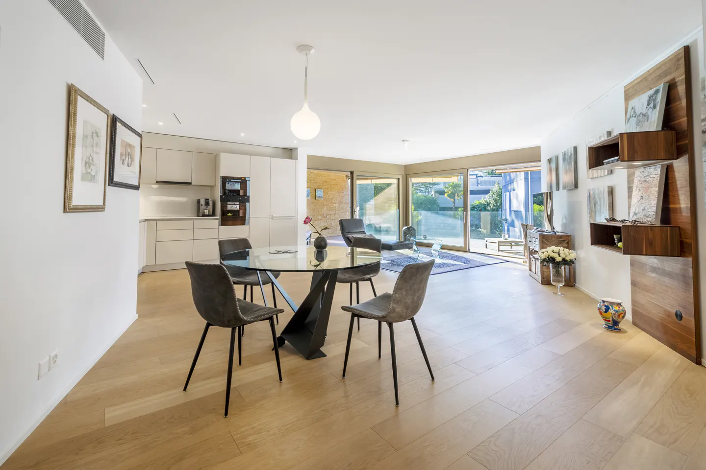 Bright, modern interior with a glass table, gray chairs, and light wood floors. Sliding glass doors lead to an outdoor area.