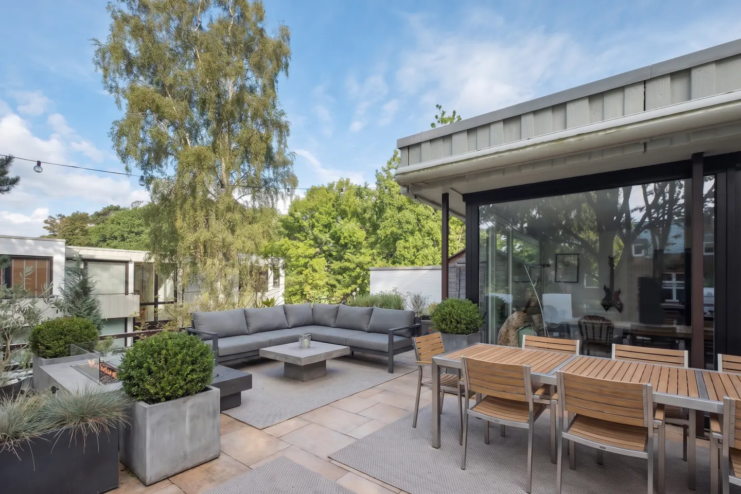 Outdoor patio with gray sectional sofa, concrete table, dining table with chairs, and planters. Trees and blue sky in background.