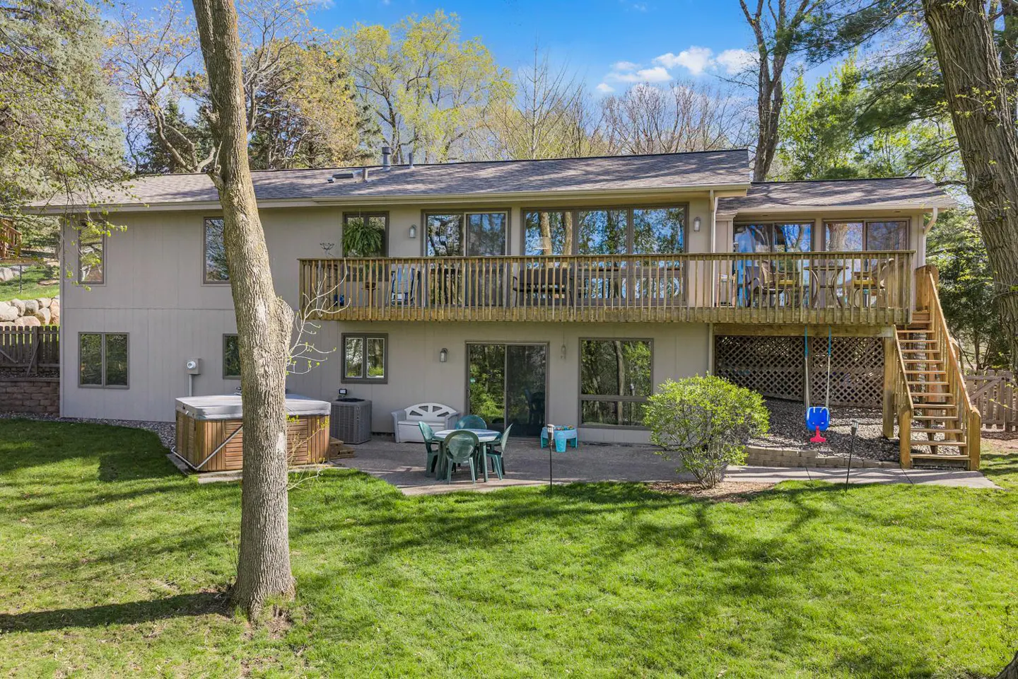 Back view of a two-story beige house with a wooden deck, stairs, patio, hot tub, and green lawn.