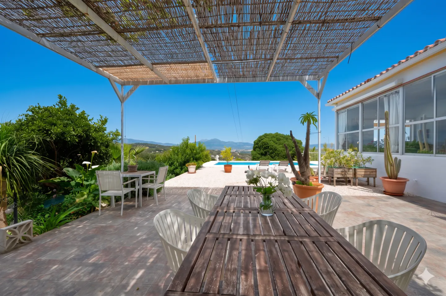 Outdoor patio with a wooden table and white chairs under a bamboo awning, overlooking a pool and mountain view.