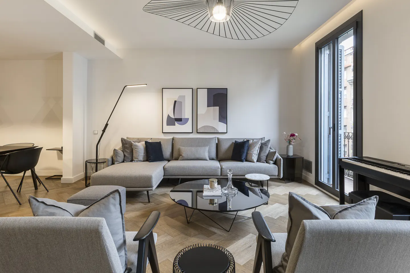 A modern living room with a gray sectional sofa, two armchairs, and a black piano near a window. A unique ceiling light fixture hangs above.