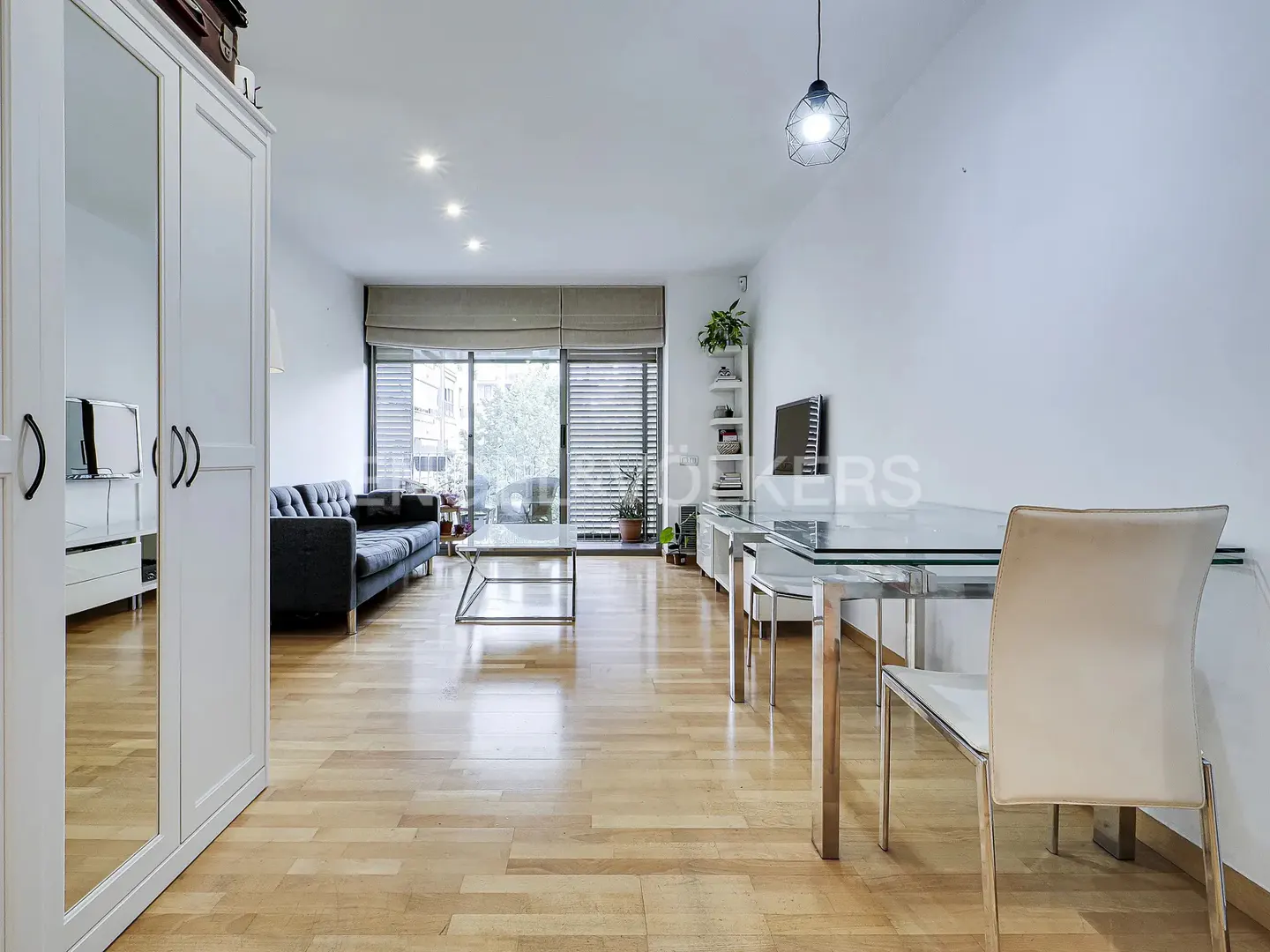Bright living room with wood floors, a black sofa, and a glass dining table with white chairs. A large window lets in natural light.