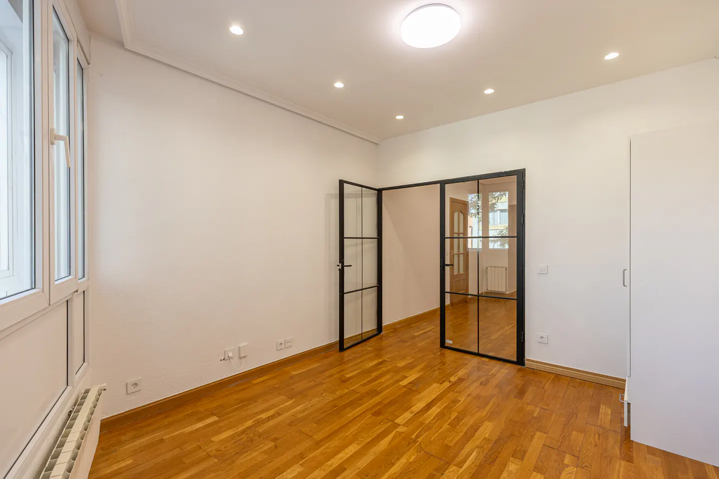 Bright, empty room with hardwood floors, white walls, and black framed glass doors leading to another room. A white window and radiator are on the left.