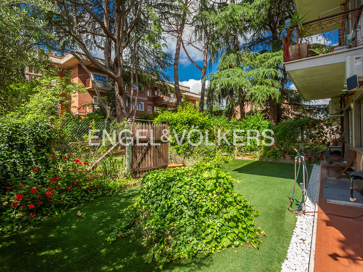 A lush green garden with trees, flowers, and a wooden gate. The Engel & Volkers logo is visible in the background.