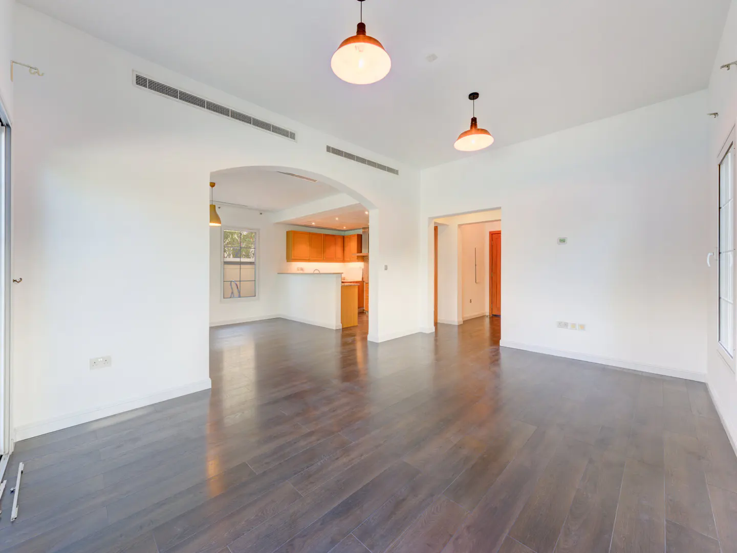 Bright, empty room with dark wood floors, white walls, and two pendant lights. Kitchen visible through arched doorway.