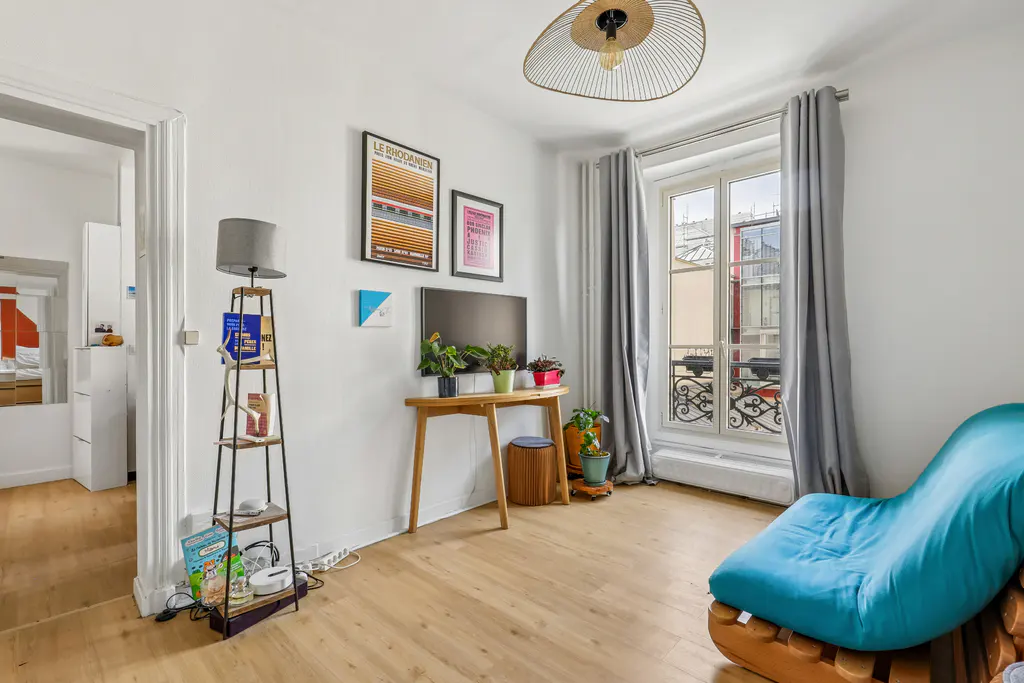 Bright living room with wood floors, white walls, and a large window with gray curtains. A blue chaise lounge sits near the window. A wooden table holds a TV and plants. Posters hang on the wall.