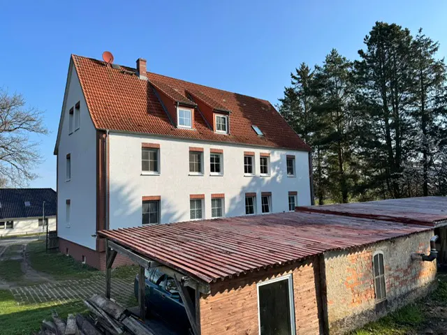 Exterior view of a three-story white building with a red tile roof and a brick outbuilding with a red corrugated metal roof.