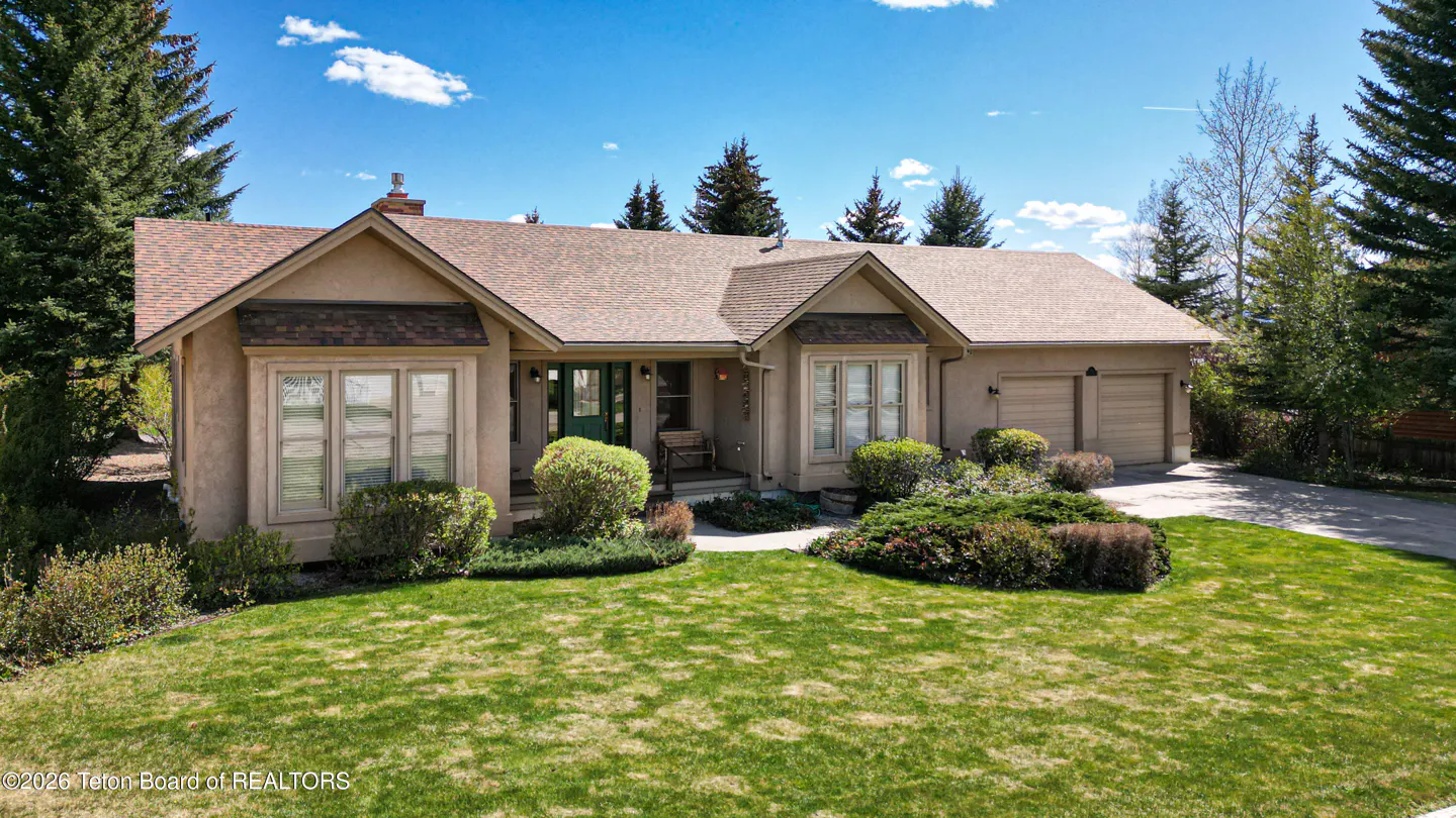 Beige single-story house with a brown roof, green door, and a two-car garage. A green lawn and bushes are in front.