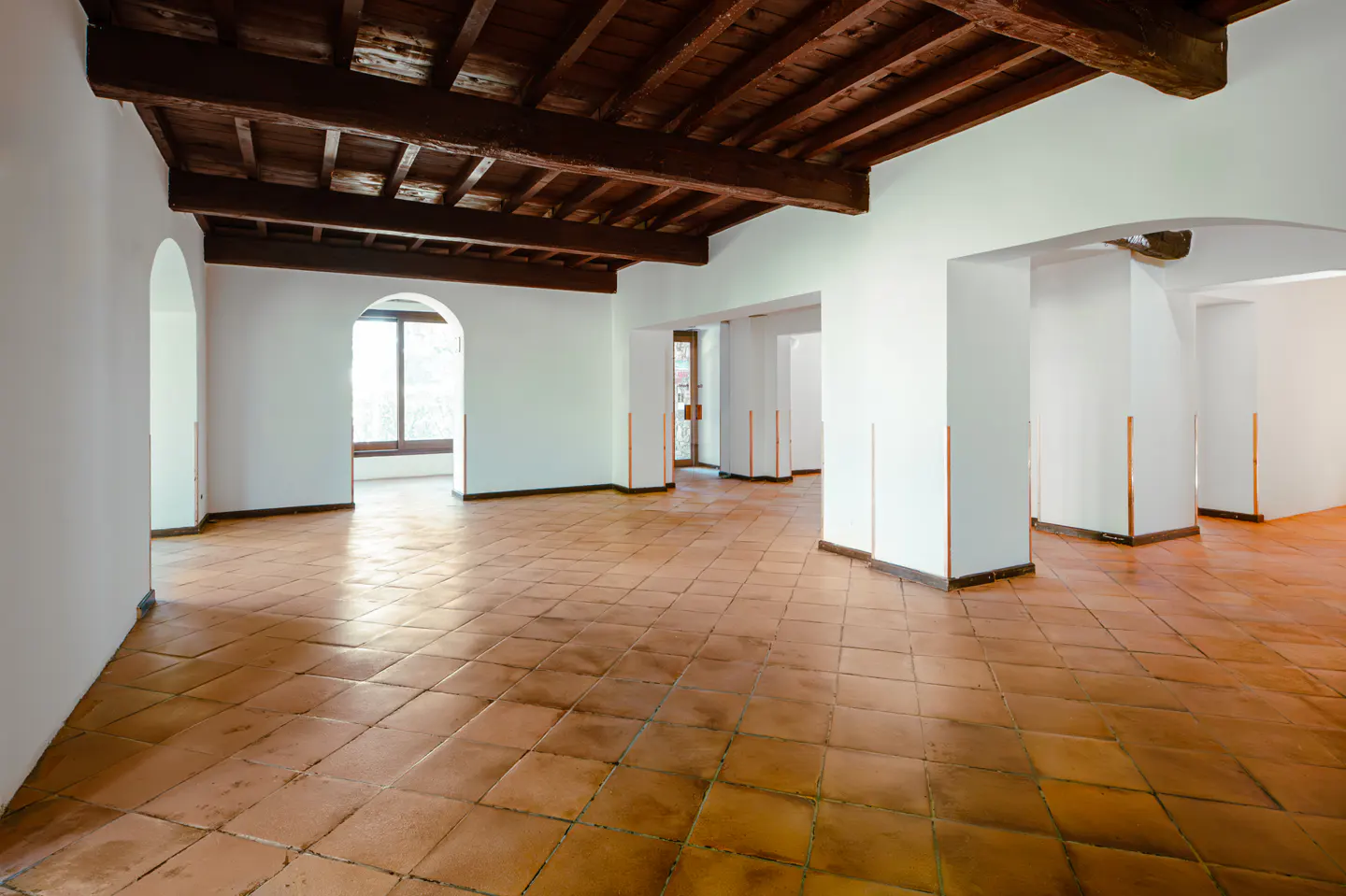 Spacious room with white walls, terracotta tile floors, and exposed dark wood ceiling beams, featuring arched doorways.