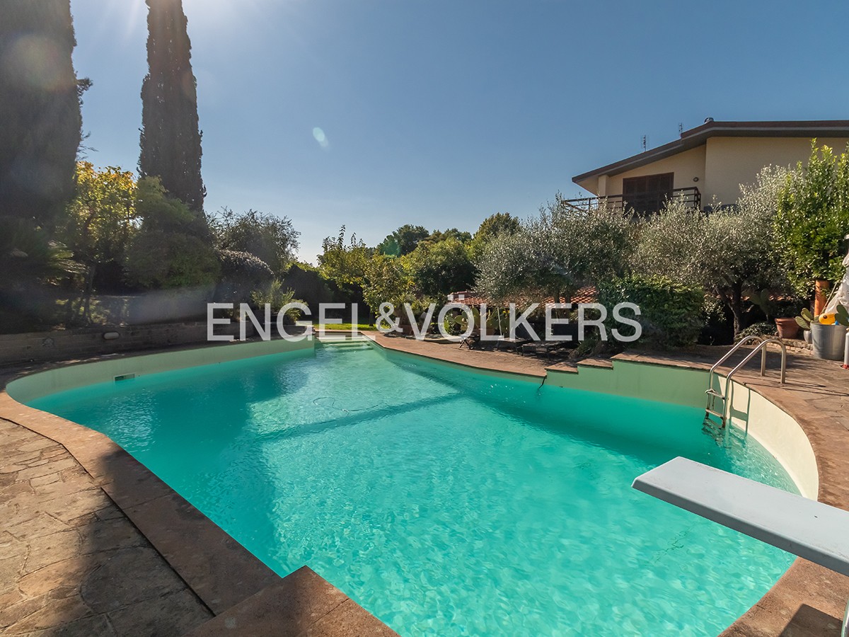 A turquoise swimming pool with a diving board and ladder, set in a sunny garden with trees and a house in the background. [AI-generated text]