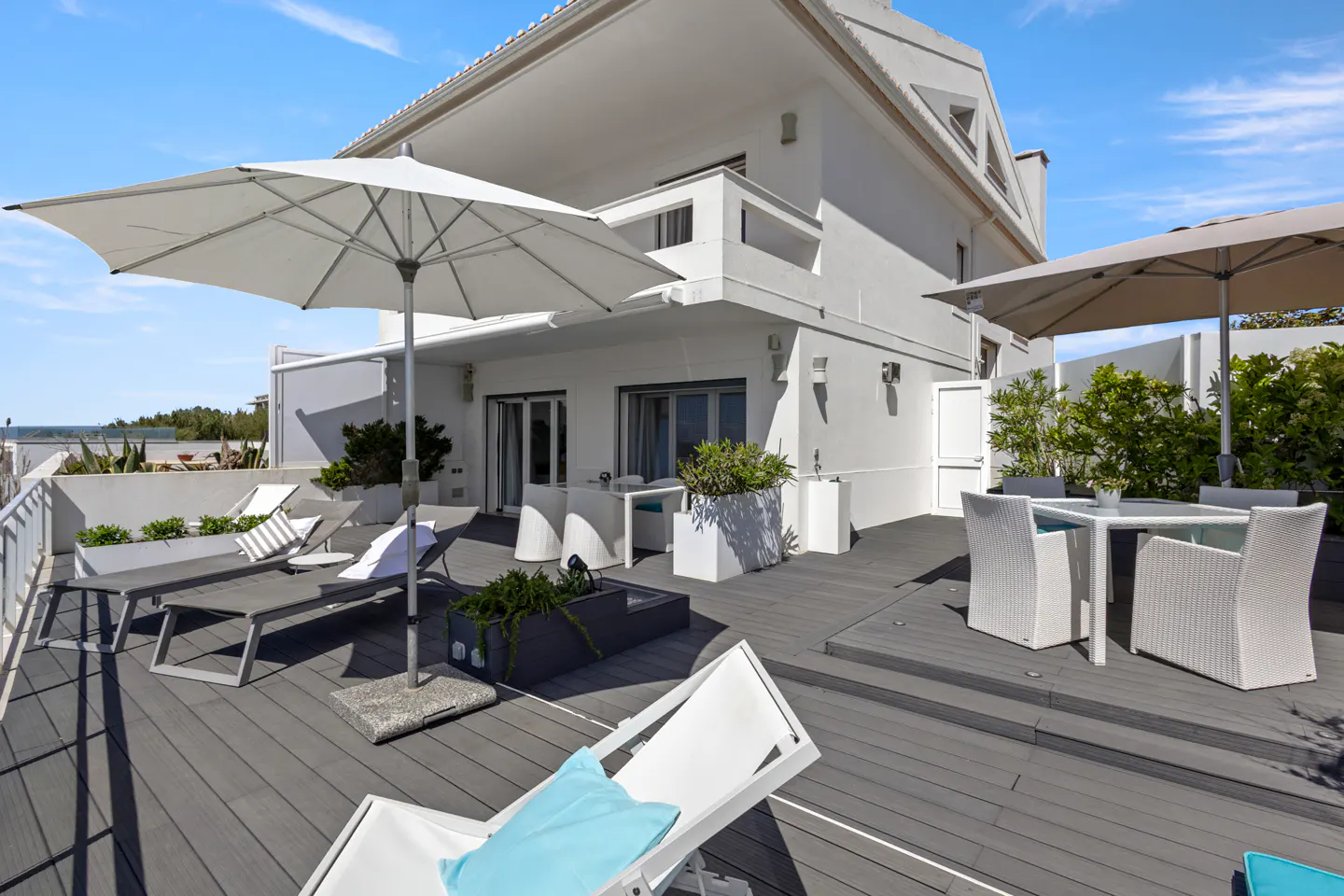 Rooftop patio with gray decking, white lounge chairs, and umbrellas. A white building is in the background under a blue sky.