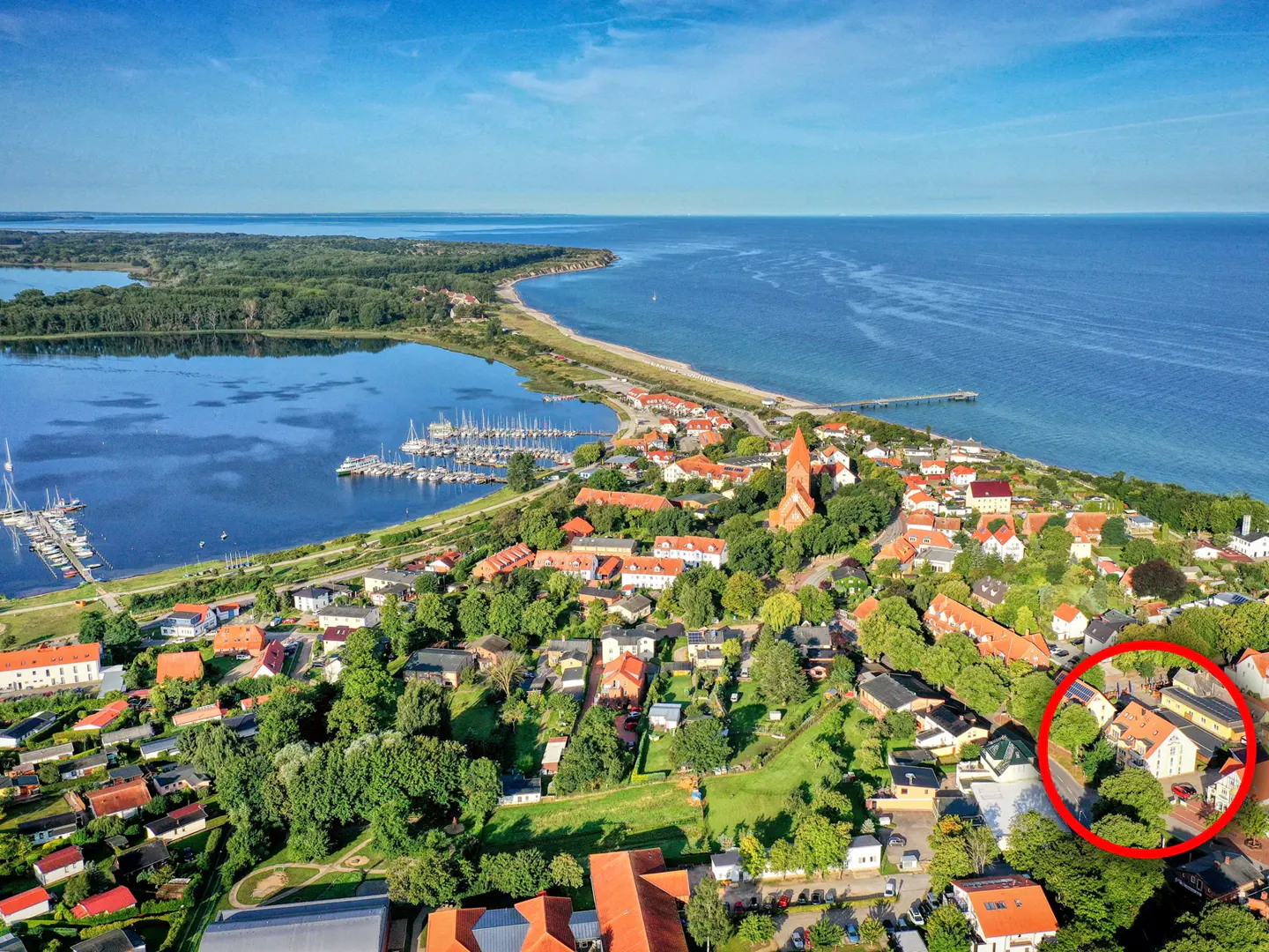 Aerial view of a coastal town with red-roofed buildings, a marina, and a church. The property is circled in red.