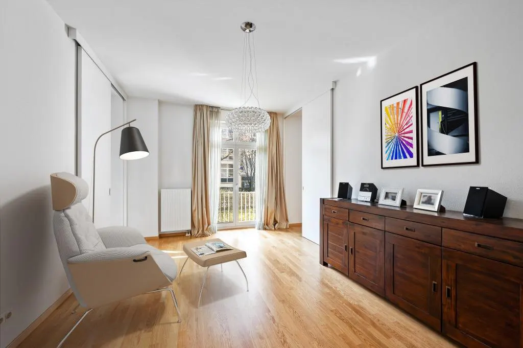 Bright living room with wood floors, white walls, and a dark wood cabinet. A white chair sits near a floor lamp and a small table with a magazine.