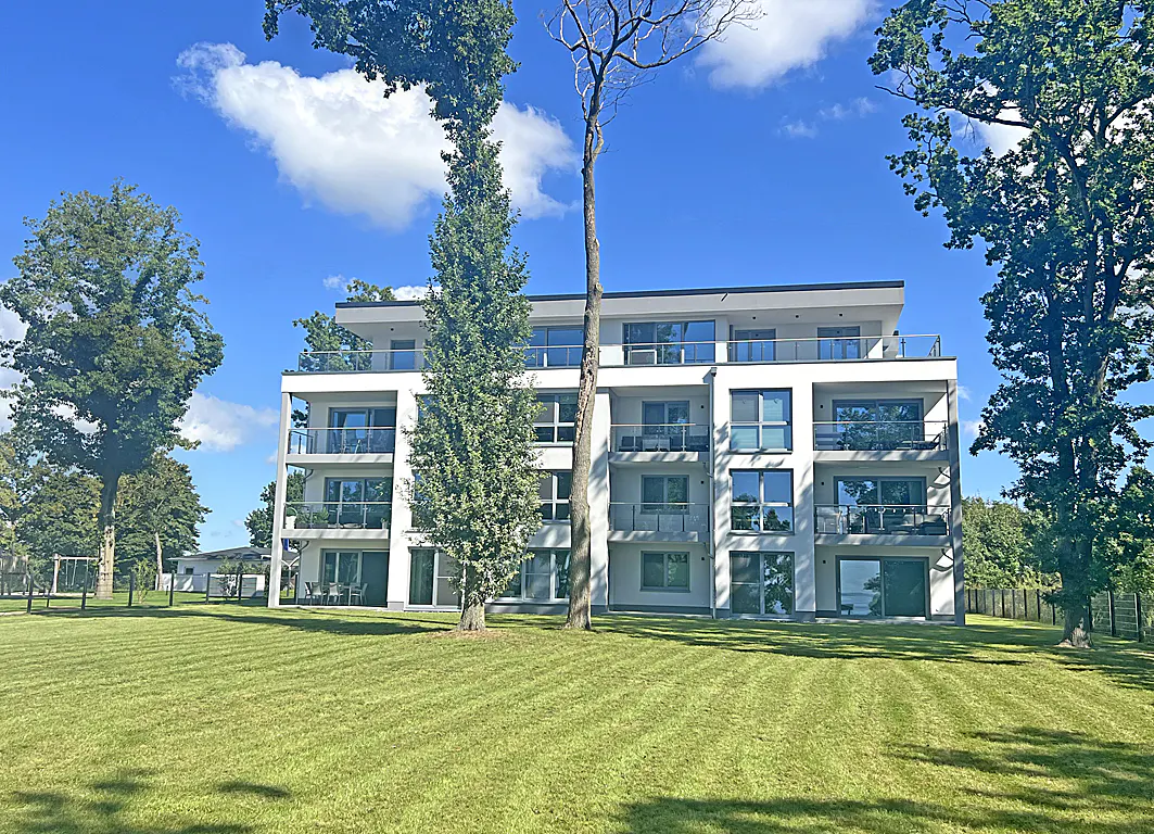 Three-story white apartment building with glass balconies, surrounded by green lawn and trees under a blue sky.
