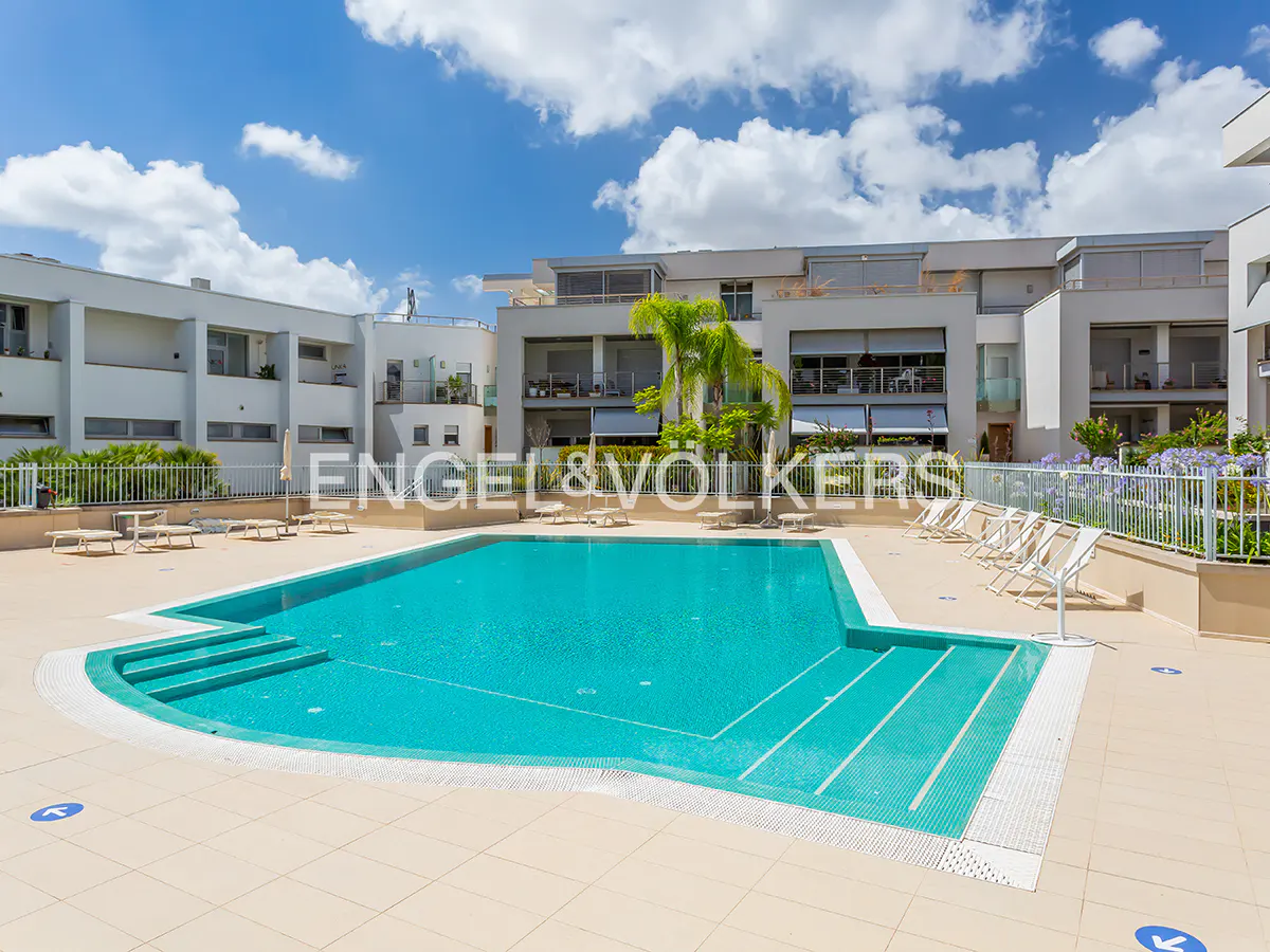 A turquoise swimming pool with steps sits in a sunny courtyard surrounded by modern apartment buildings and palm trees.