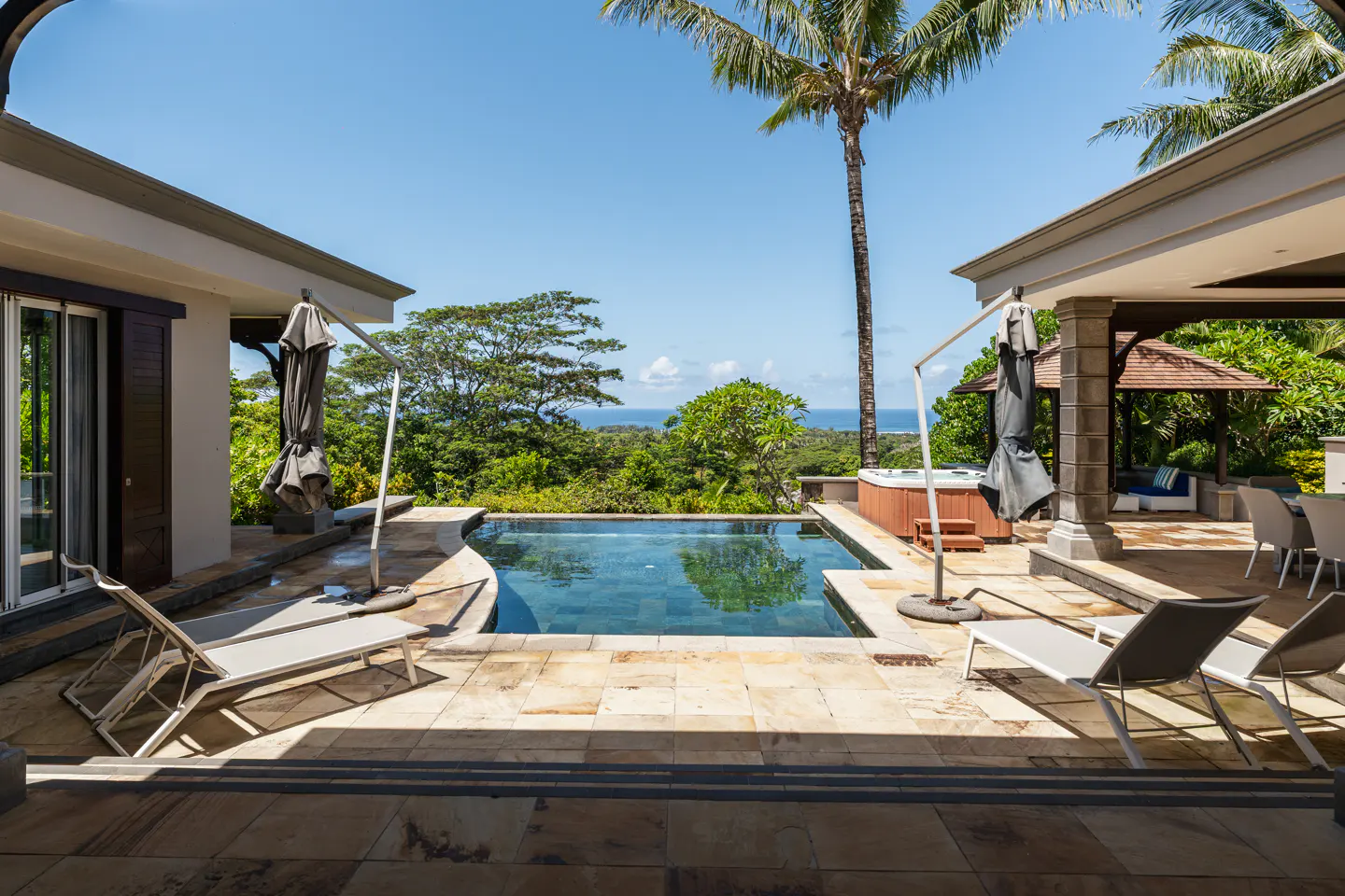 Luxury villa with pool overlooking lush greenery and ocean. Lounge chairs and umbrellas surround the pool. Blue sky above.