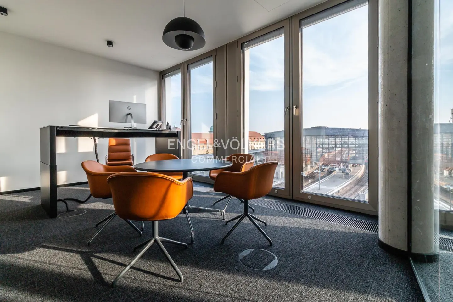 Office space with a round table, orange chairs, and a desk with a computer. Large windows offer a city view.