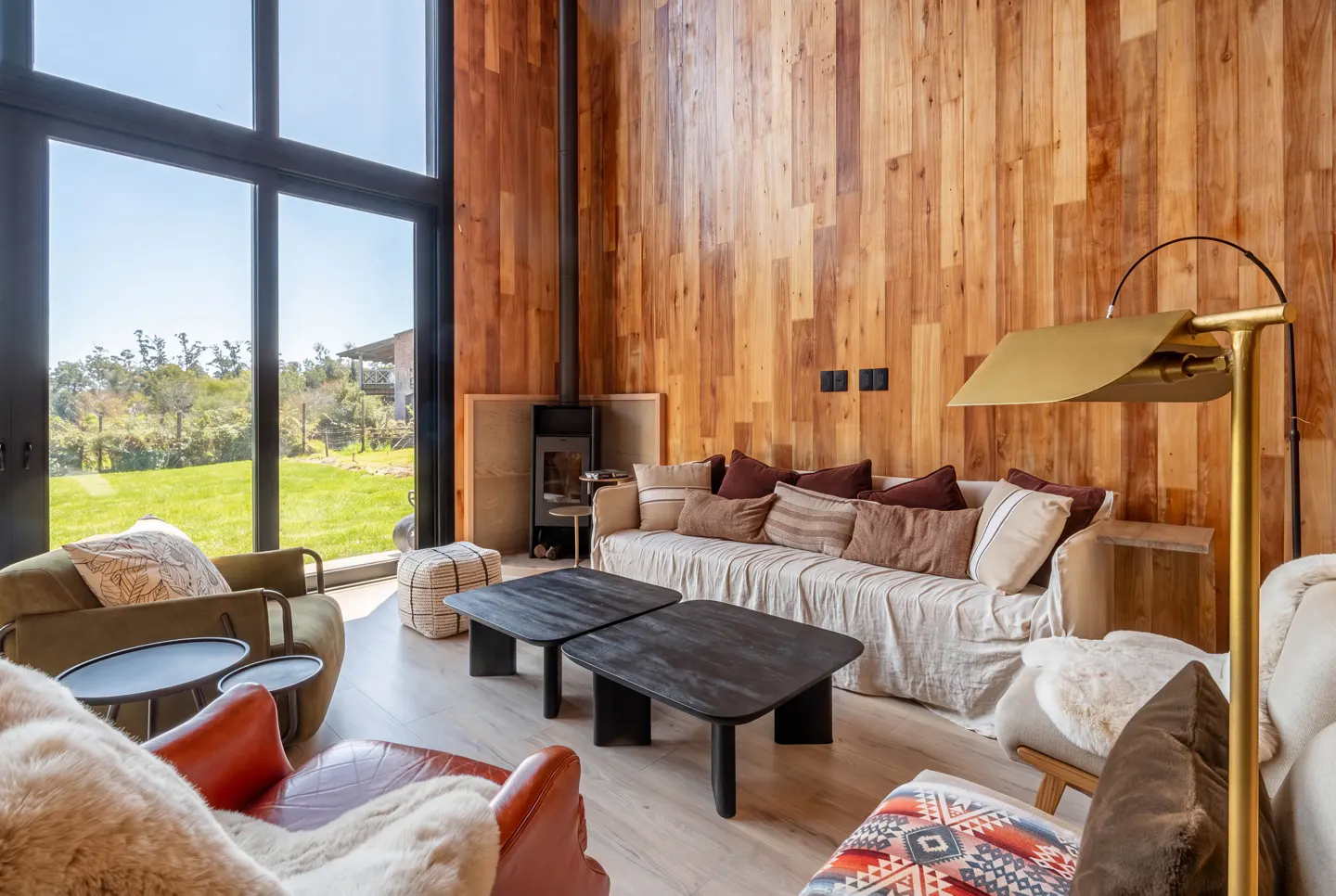 Modern living room with wood-paneled walls, floor-to-ceiling windows, a fireplace, and cozy seating facing a green yard.
