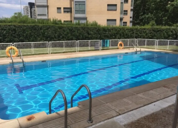 Outdoor pool with blue water and lane lines, surrounded by a white fence, green grass, and a hedge. Apartment buildings are in the background.