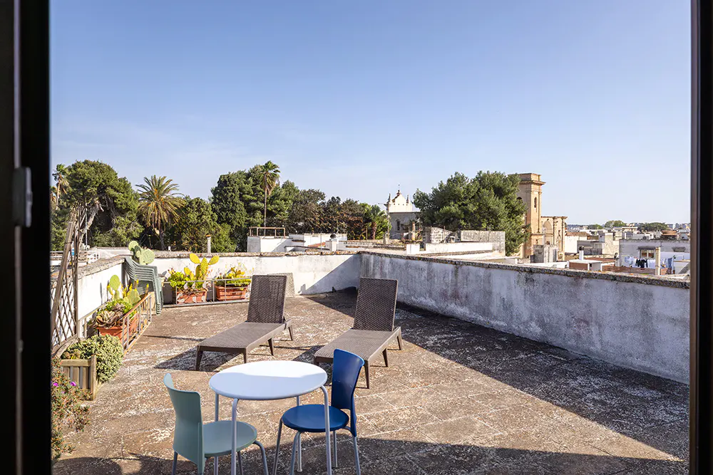 Rooftop patio with two lounge chairs, a round table, and three chairs. Cityscape and blue sky in the background.