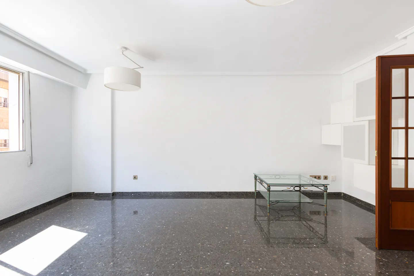 Bright, empty room with white walls, a dark speckled floor, and a glass table. A window and a wood-framed door are visible.
