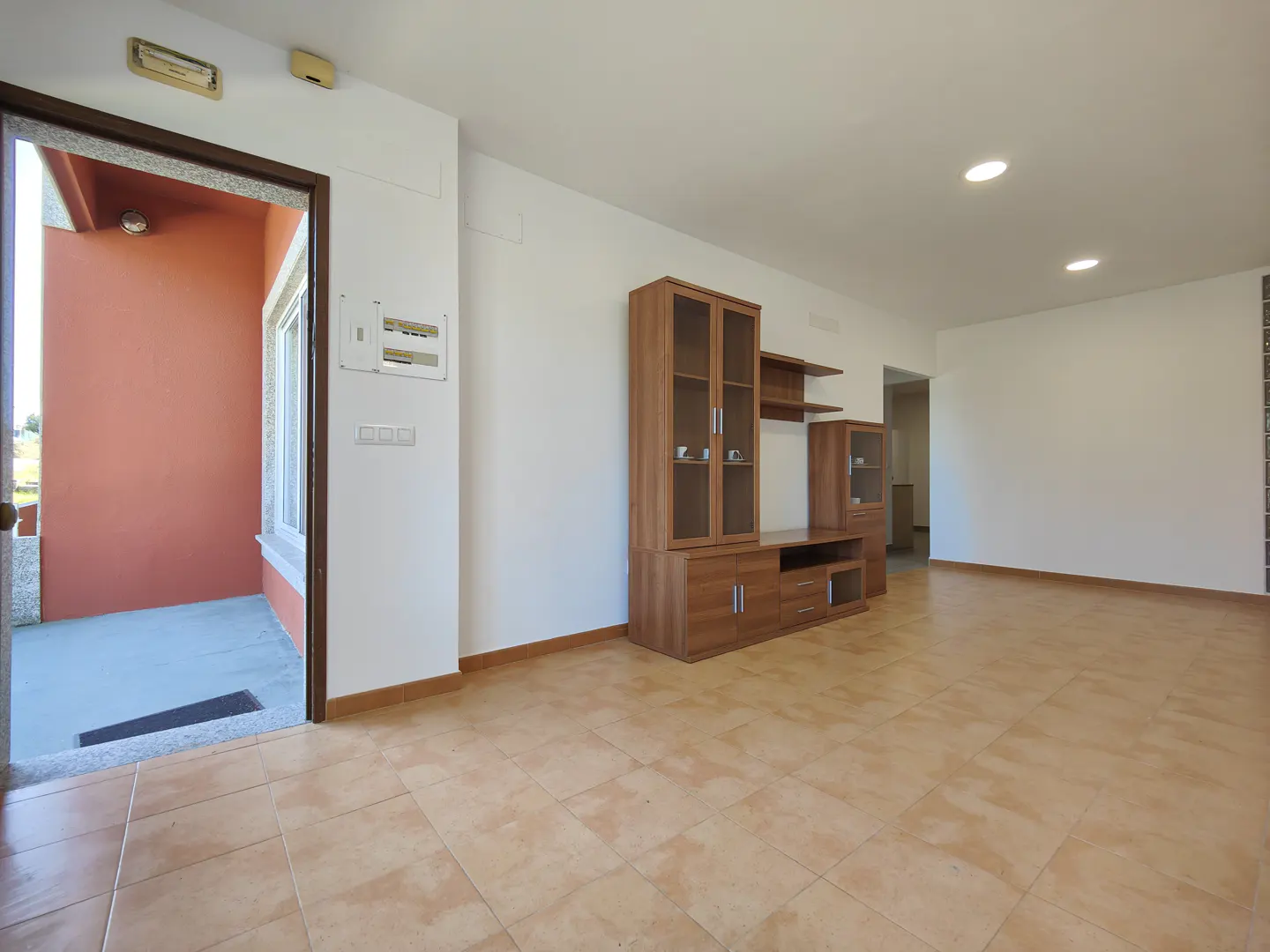 A bright, empty living room with tile floors, white walls, and a wooden entertainment center. An open doorway leads to an orange-walled porch.