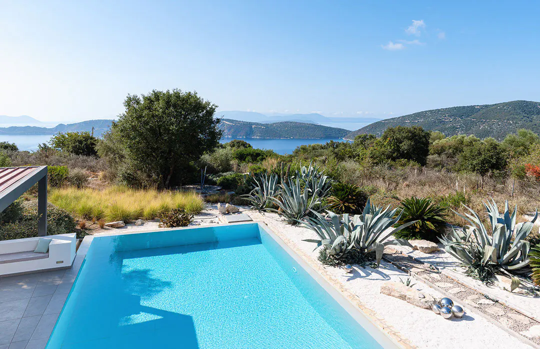 A bright blue pool is surrounded by white gravel, agave plants, and greenery, with a view of the ocean and islands in the background.