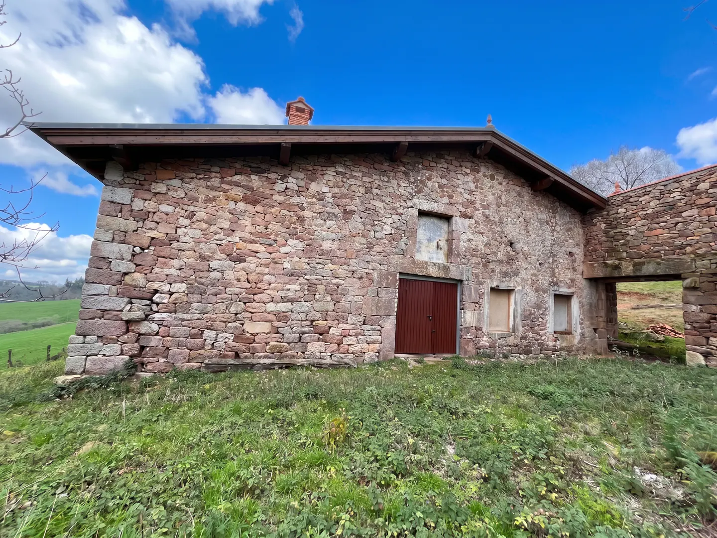 Exterior view of a stone barn with a brown door, set against a blue sky with clouds and a green field.