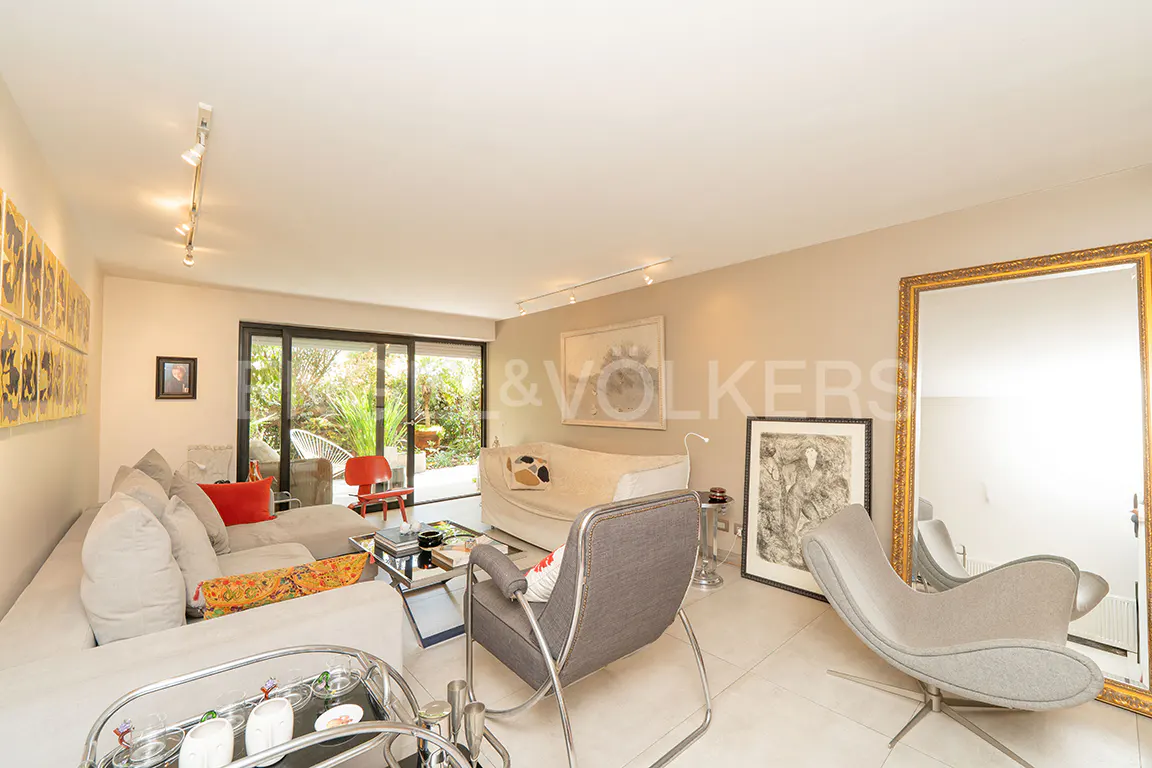 Bright living room with a large sectional sofa, gray chairs, and a gold-framed mirror. Sliding glass doors lead to a lush garden.