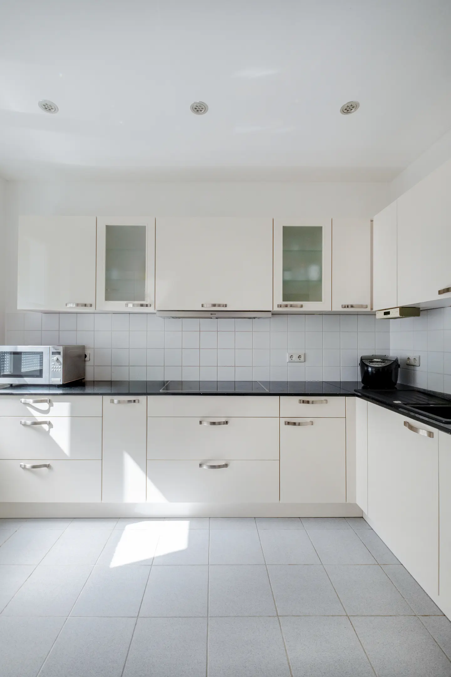 Bright, modern kitchen with white cabinets, black countertops, and gray tiled floor. Appliances include a microwave and toaster.