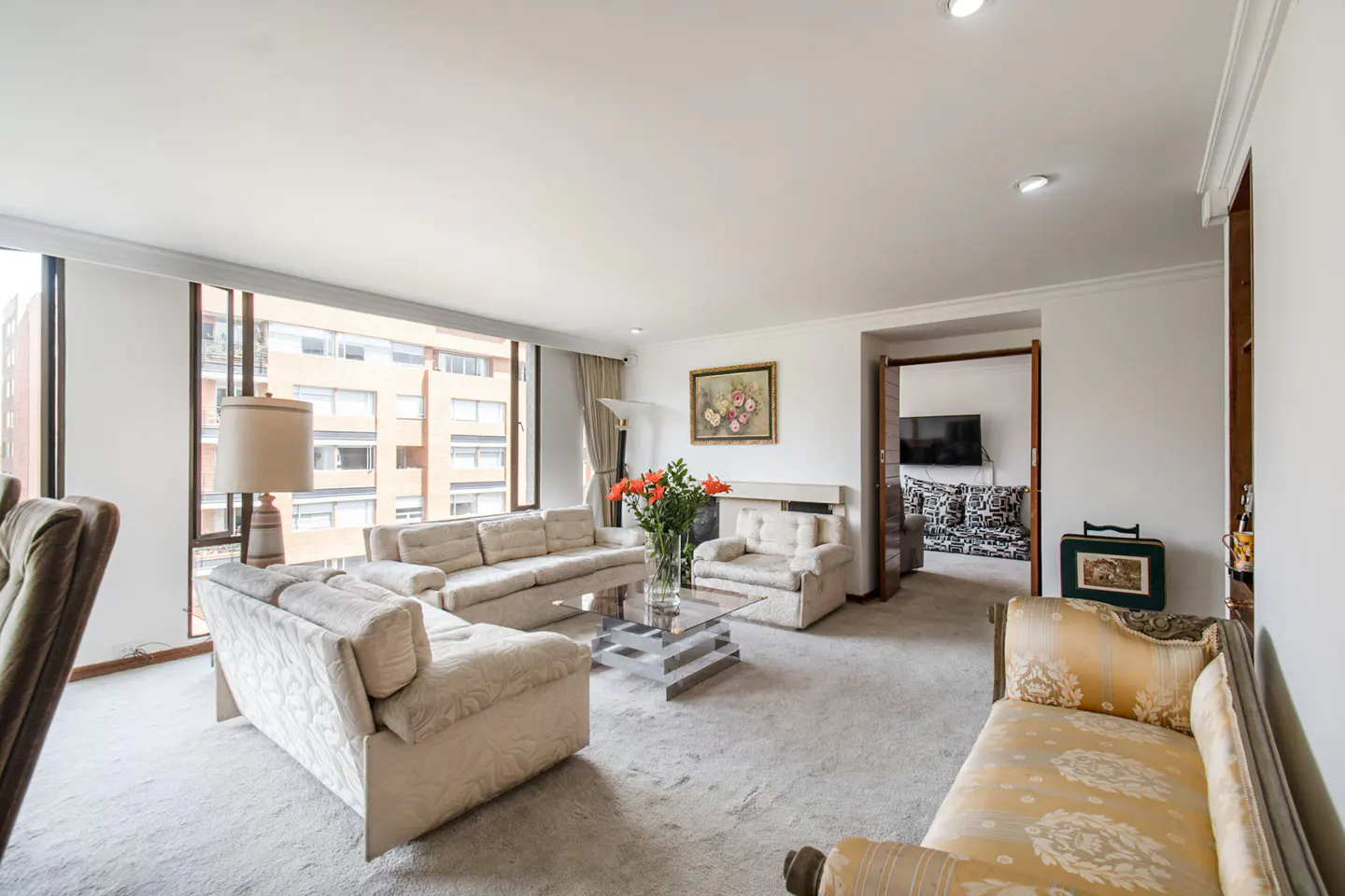 Bright living room with white sofas, glass table, and orange flowers. Large windows overlook city buildings. A doorway leads to another room.