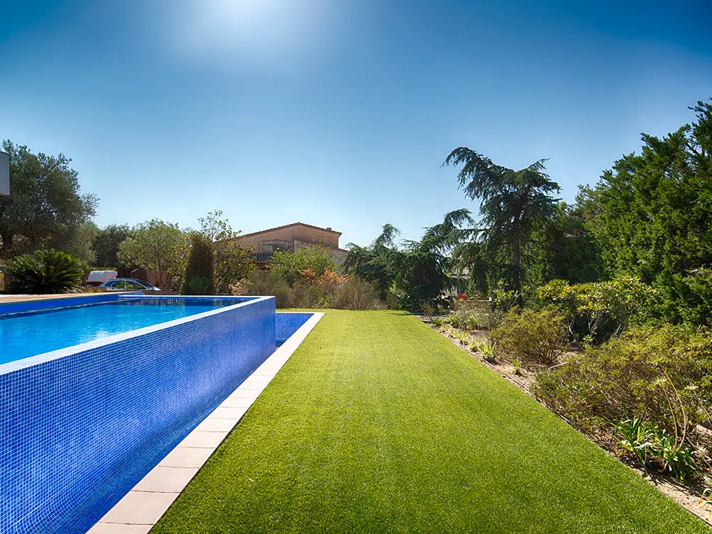 A bright blue tiled pool sits next to a green lawn under a clear blue sky. Trees and a building are in the background.